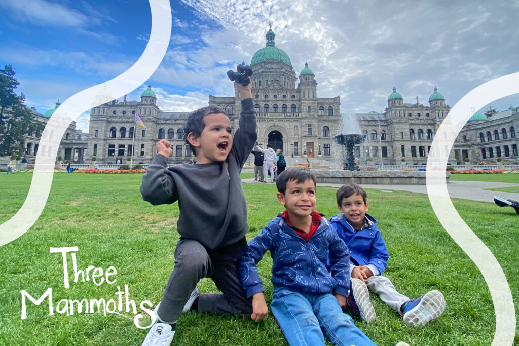 Three kids laughing and playing in front of the historic Parliament Buildings in Victoria, BC, with the iconic architecture and lush green lawn creating a picturesque backdrop